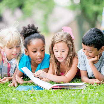 A group of kids reading a book together in the grass.