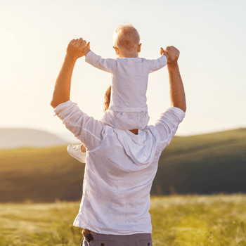 A dad with a little boy on his shoulders in a grassy field.