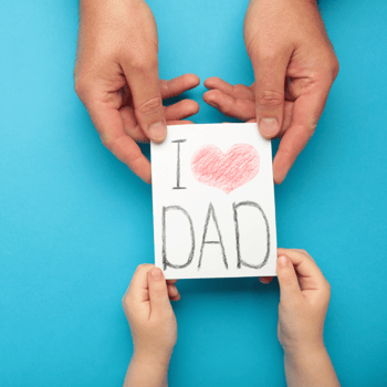 A card that says "I love dad" given by a little boys hands to his dads hands.