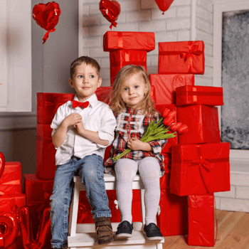 A little boy and girl sitting on a bench surrounded by red boxes and heart shaped balloons.