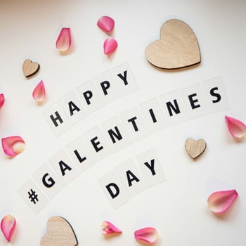 Letters that say "Happy #Galentines Day" surrounded by rose petals and wooden hearts.