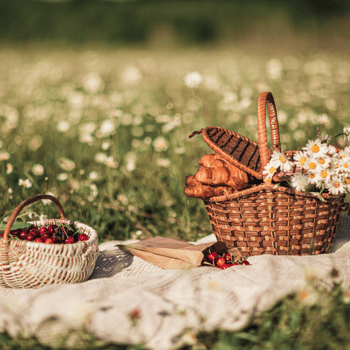 A cute picnic basket surrounded by flowers on a cream colored blanket.