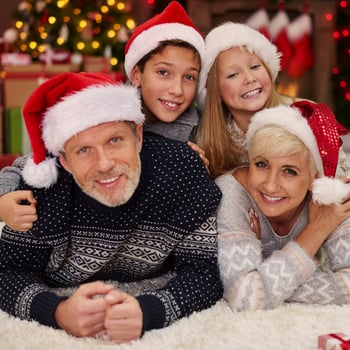 A family taking a photo together with Santa hats in front of their Christmas tree.