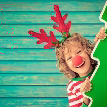 A little kid with a red nose and antlers on hiding behind a cardboard tree.