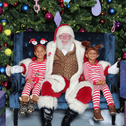 Photo of siblings with Santa. One wearing reindeer antlers and another wearing a headband with little Santas. Both are in matching pjs.