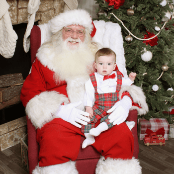 Baby boy dressed in plaid overalls sitting with Santa.