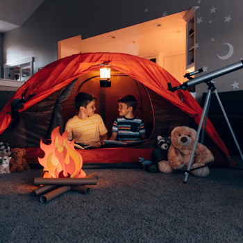 Two boys in a red tent in a living room. Set up with a fake fire and telescope.