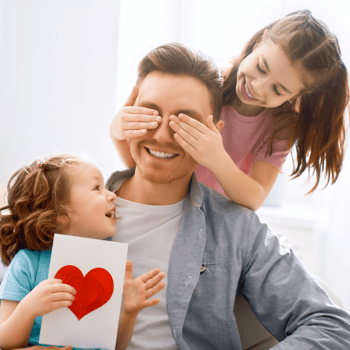 Two little girls covering dads eyes with a handmade card.