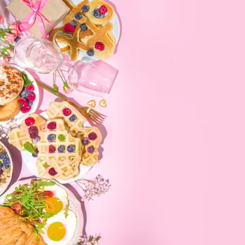 Brunch food with X and O pancakes, raspberries, and more on a pink background.