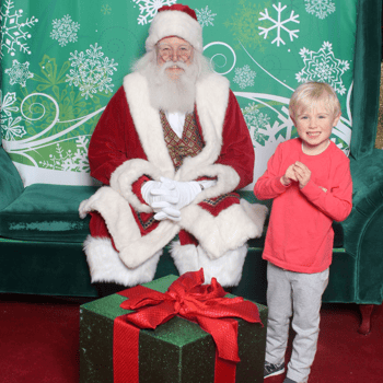 Little boy standing next to Santa with his hands clasped together and a present on the floor.