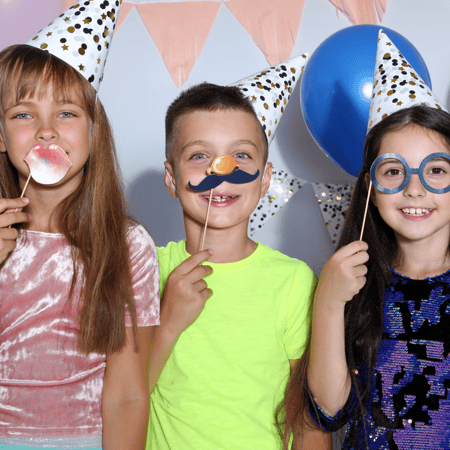 Three kids wearing confetti party hats and using fun props for a photo booth.