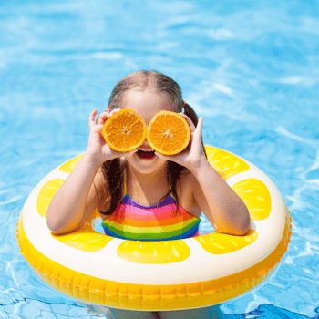 A little girl in an orange floaty ring holding up orange slices to her eyes.