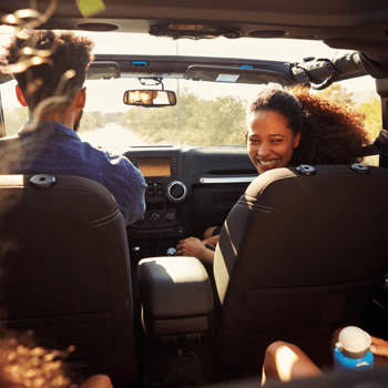 A mom smiling at her kids in the backseat from the passenger seat of a car while on a road trip.e