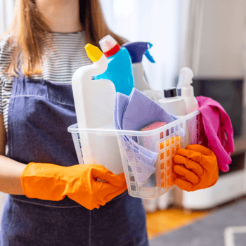 A woman holding a basket of cleaning supplies with orange gloves on.