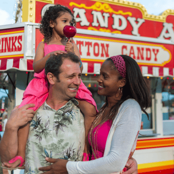 A little girl eating a candy apply on her dads shoulders and mom looking at them at the fair.