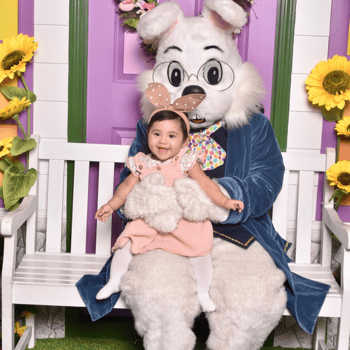 A little girl in a pink dress with tan bunny ears sitting with the Easter bunny.