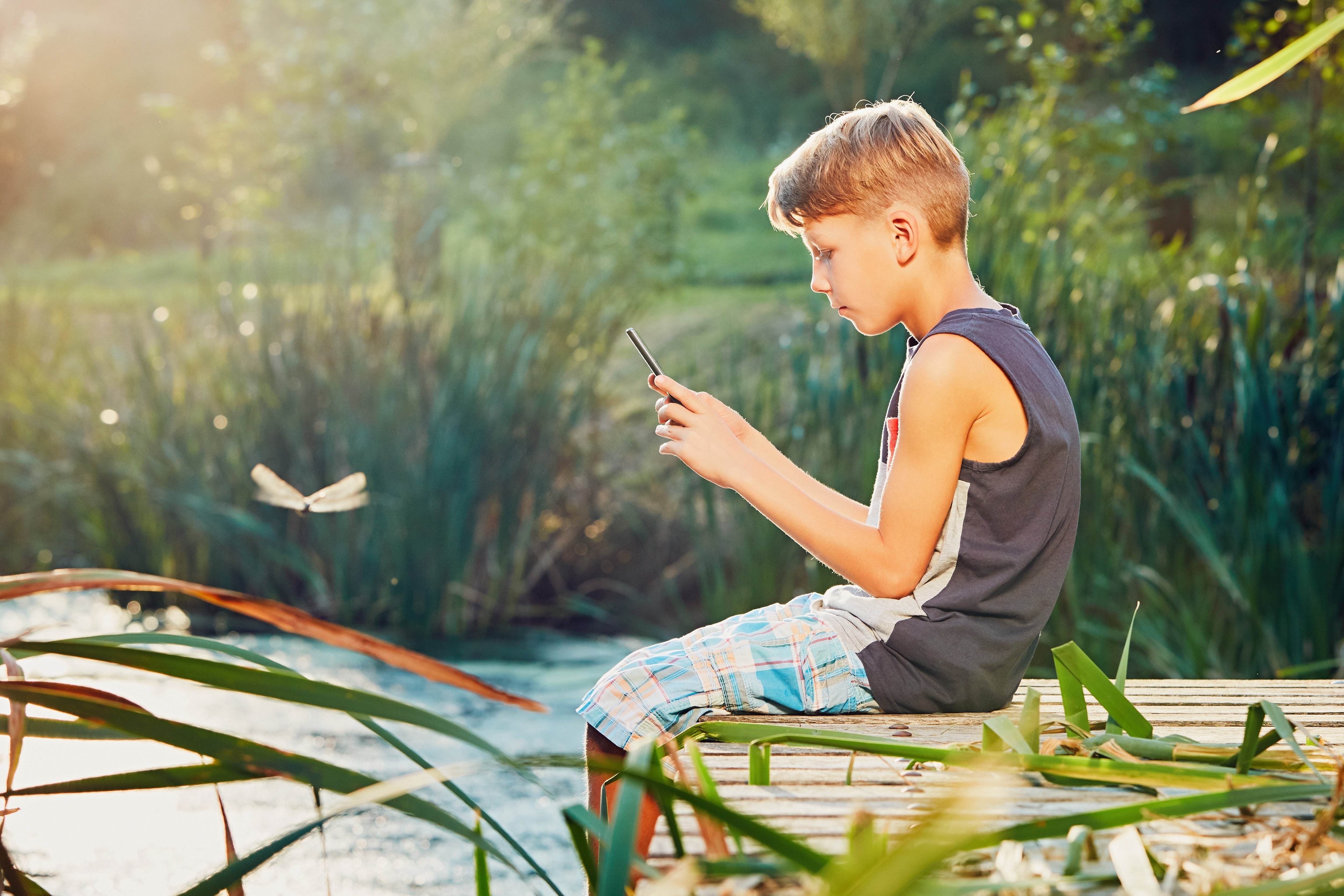 boy taking photo of dragonfly with phone seated on a dock sitting by the water summer day