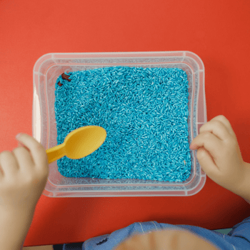 A container of blue rice and a toddler with a spoon to play with.