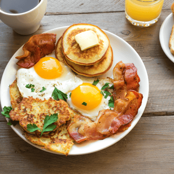 A plate of eggs, bacon, and pancakes on a wood table with orange juice and coffee.