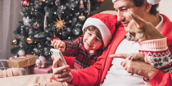 child and parent and dog all wearing santa hats and festive attire while pointing and looking at a cell phone with a christmas tree in the background