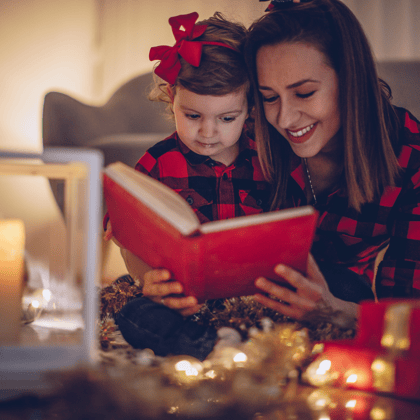 A mom and little girl in red Christmas pajamas reading a book together.