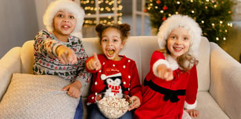 three children sitting on a couch watching a Christmas movie and eating popcorn while wearing festive attire and Santa hats