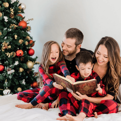 Family gathered in matching pj's and reading a book next to a Christmas tree.