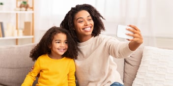 mother and daughter sitting on sofa couch smiling and taking selfie with a mobile device