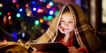 child reading on a tablet under a blanket with christmas tree in the background with the lights off